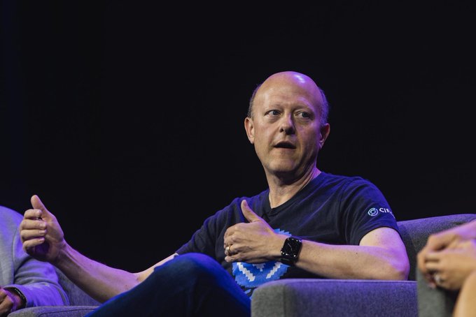 A bald man in a dark t-shirt speaking on a stage with purple lighting, gesturing with his hands while seated.