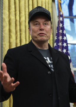 A photo of Elon Musk wearing a black 'Make America Healthy Again' cap and a dark blazer over a t-shirt, speaking and gesturing with his hand in what appears to be the Oval Office.