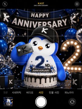 A photo taken through a smartphone camera showing a 'HAPPY K ANNIVERSARY' 2nd anniversary celebration featuring a penguin mascot wearing a black cap with a cake in front of it.