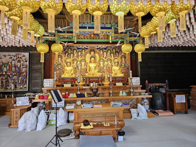 A Buddhist temple interior features golden Buddha statues on an altar beneath rows of ornate golden lanterns and hanging prayer scrolls, with Korean text visible on signs including "통도" and English notices. The scene of spiritual reverence and timeless tradition contrasts with the post's speculative "$500 by 2030" price prediction for $SATL, juxtaposing ancient serenity against modern crypto ambition.