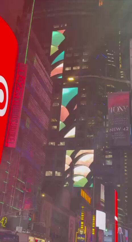 The image shows a city street at night with tall buildings lining both sides. The buildings are illuminated by colorful lights that create a vibrant atmosphere. In the foreground of the photo, there's an advertisement for a new product called "Floki". This advertisement is prominently displayed on one of the buildings and stands out against the dark background. The presence of this advertisement suggests that the city has a thriving business environment or that it is known for its unique and eye-catching advertisements.