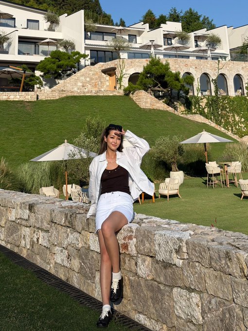 A woman poses on a stone wall at an upscale outdoor resort with terraced gardens and patio umbrellas behind her, complementing the post's announcement of her first day working at Polymarket in Singapore.