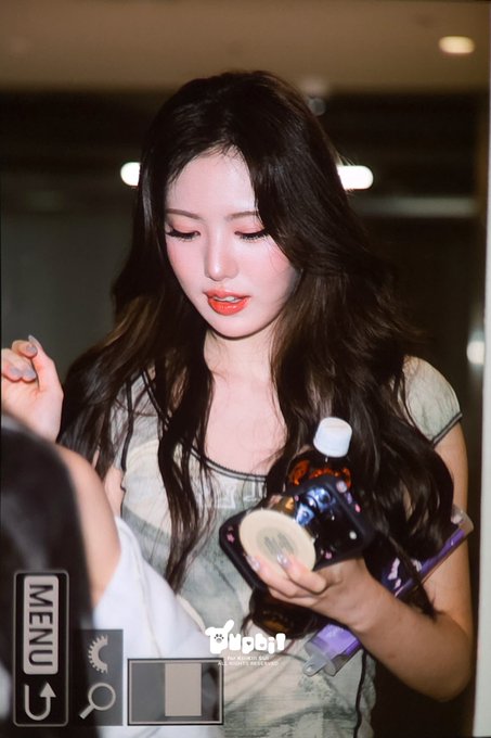 A photo of a young woman with long dark wavy hair and orange lipstick, holding a bottle, with 'MENU' and 'U+' camera UI elements overlaid and 'Mobil' watermark visible at the bottom.