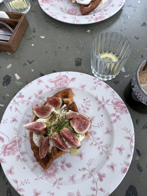 A decorative plate featuring toast topped with fresh fig slices, cream or cheese, and herbs, accompanied by a glass of lemon water and a coffee cup on a terrazzo surface.