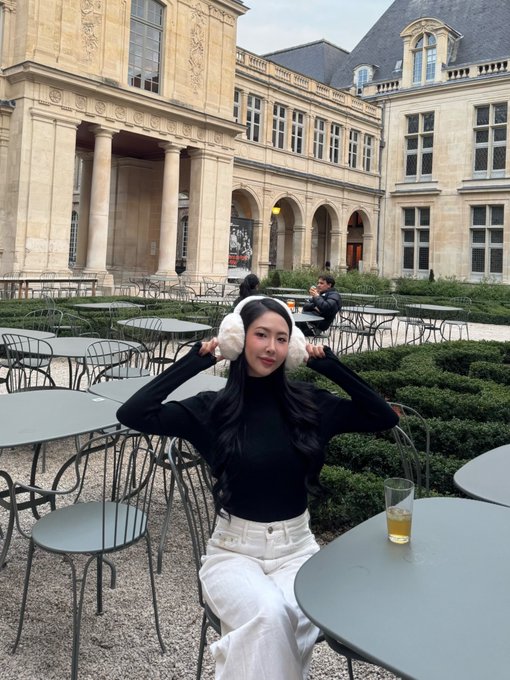A woman with long dark hair and white earmuffs sits at an outdoor cafe in front of a grand classical building, wearing a black turtleneck and white pants.