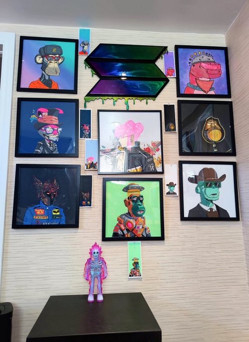 An interior room display featuring a gallery wall of framed NFT-style digital artworks depicting various Bored Ape-style characters, with a glowing pink and purple figurine displayed on a black pedestal below.