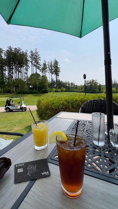 The image shows a table with two drinks, one of which has a lemon slice floating in it. The table also has a credit card placed next to the drinks. In the background, there are trees and a golf cart visible, suggesting that this is an outdoor setting or a golf course.