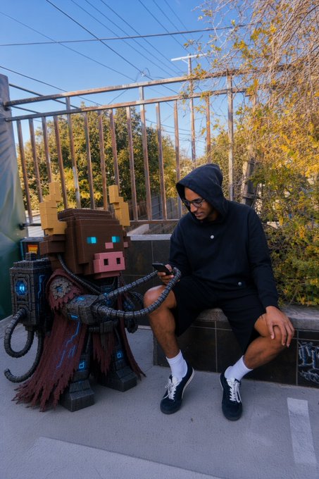 A man in a black hoodie and shorts sits on a ledge next to a large LEGO-style sculpture of a mechanical bull, looking at his phone.