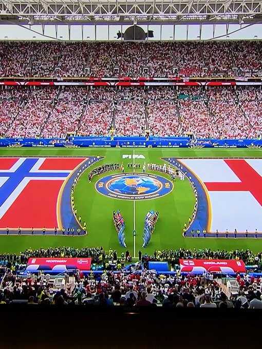 A wide shot of a packed soccer stadium during a pre-match ceremony displays giant 'NORWAY' and 'ENGLAND' flags on the pitch flanking a UEFA EURO 2024 center circle logo, with the teams lined up in the middle.