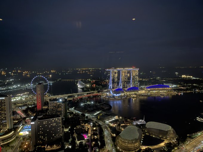 A nighttime aerial cityscape of Singapore, prominently featuring the illuminated Marina Bay Sands hotel, the Singapore Flyer, and the ArtScience Museum.