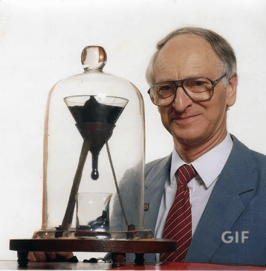 A photograph of an older man in a suit and glasses standing next to the famous pitch drop experiment, with 'GIF' watermarked in the corner.