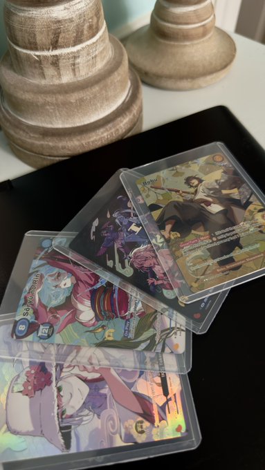 A collection of anime-themed trading cards in protective sleeves is fanned out on a dark surface, with decorative wooden candle holders visible in the background.