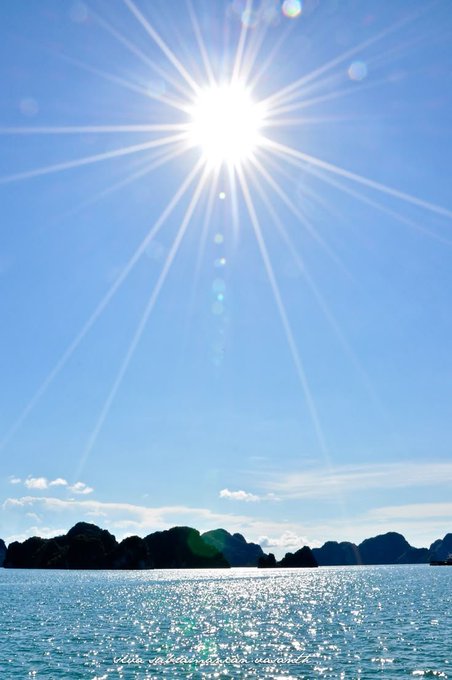 A bright sun shines over a calm blue sea with rocky islands on the horizon, casting a sparkling reflection on the water.