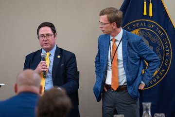 Two men in suits at an event with a 'STATE OF NEBRASKA' flag visible; the man on the left speaks into a microphone while the man on the right wears a blue blazer and orange tie.