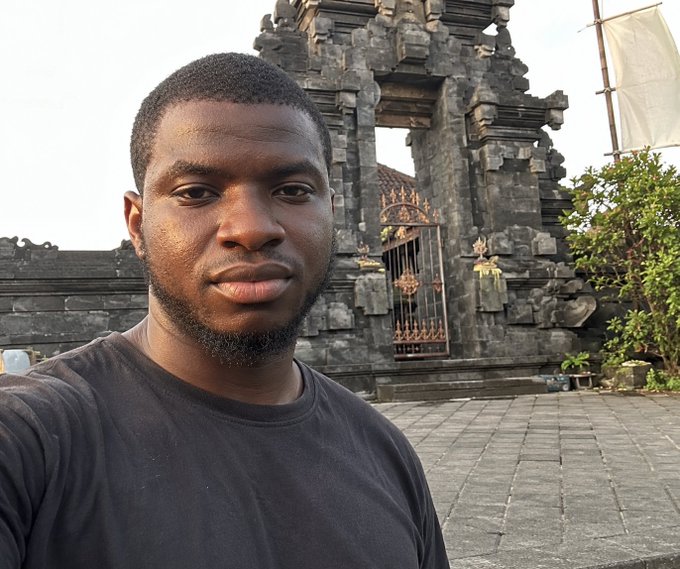 A selfie of a young man wearing a black t-shirt, taken in front of an ornate Balinese temple gate with traditional stone carvings and a metal gate.