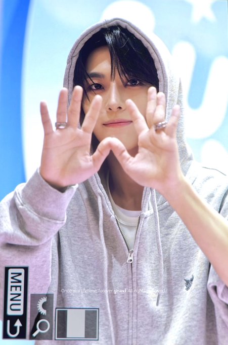 A young man with dark hair, wearing a light gray hooded zip-up sweatshirt, holds his hands up in front of the camera forming a heart shape with his fingers. The image appears to be a fan-taken photo, with "MENU" text in the bottom left corner and the watermark "Once in a Lifetime, forever yours! All rights reserved" overlaid on the image. Based on the hashtags (#XINLONG, #鑫隆, #씬롱, #ALPHADRIVEONE, #ALD1), this appears to be a fan photo of Xinlong, a member of the K-pop group ALD1.