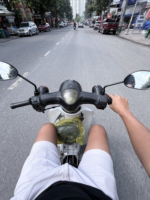 A rider's perspective from atop a scooter navigating a bustling street in Vietnam (with a "NHA THUOC TAY" pharmacy sign visible on the right) while a helmet rests on the fuel tank between their legs. The image's open road ahead symbolizes the "stay calm and keep moving" mindset of the post's crypto investment message—buying low requires steady nerves, not emotional reactions when markets dip.