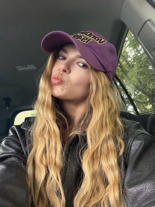A selfie of a woman with long blonde hair making a kissing face, wearing a purple 'Vegas Golden Knights' cap and a black leather jacket inside a car.