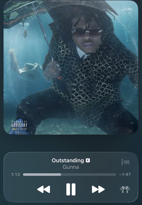 A music player showing the song "Outstanding" by Gunna with parental advisory explicit content, displayed over an album artwork featuring a man in a patterned jacket holding an umbrella underwater.
