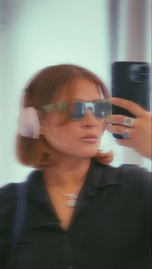 A stylish woman with a short bob taking a mirror selfie, wearing green-tinted sunglasses, pink headphones, and multiple rings.