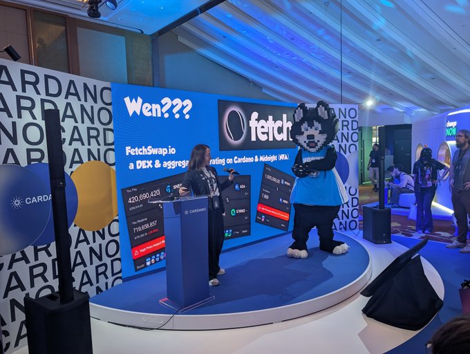 A trade show or expo booth for "fetch" featuring a presenter in a leather jacket on stage next to a mascot in a blue shirt, with a large screen displaying "Wen??? FetchSwap.io a DEX & aggregating on Cardano & Midnight (WFA)."