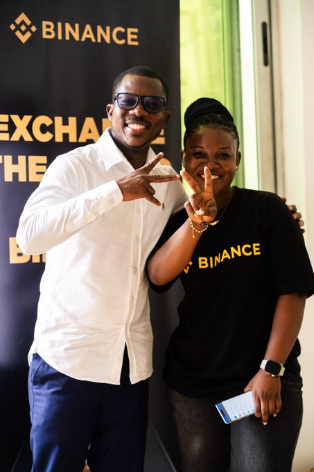 A man in a white shirt and sunglasses and a woman in a black Binance t-shirt pose together with peace signs in front of a Binance event backdrop.