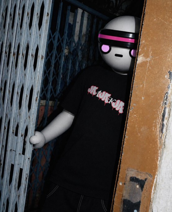 A mannequin wearing a black t-shirt with pink gothic-style 'JAKRUA' text, standing in a doorway next to a metal gate.