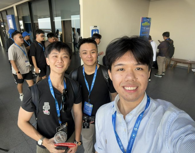 A selfie taken at Coinfest Asia shows three smiling attendees wearing event lanyards and badges, with other participants and event signage visible in the background, capturing the casual networking moment between familiar faces at the conference.