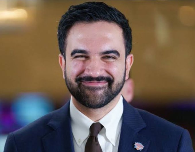 A headshot of a smiling man with dark hair and a full beard, wearing a dark suit, white shirt, and brown tie with a small pin on the lapel.