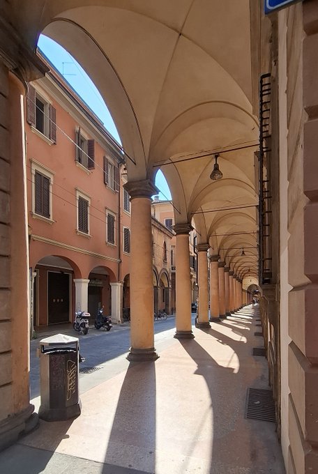 A view down a covered arched walkway with columns, leading to a sunlit Italian street lined with peach-colored buildings.