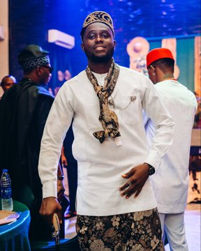 A man wearing a white traditional outfit with a patterned cap and matching scarf stands at what appears to be a formal event, with other attendees visible in the background.
