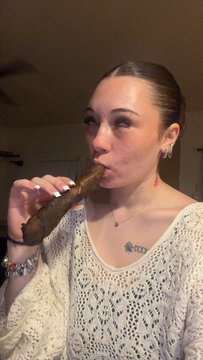 A young woman with a sleek ponytail and earrings poses indoors while holding a brown cigar near her face, wearing a white crocheted top.