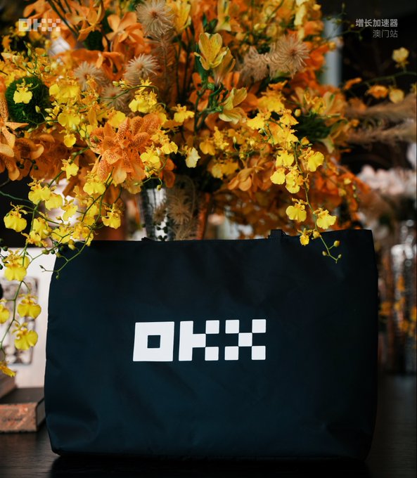 A dark blue tote bag with the white 'OKX' cryptocurrency exchange logo sits on a surface in front of an arrangement of yellow and orange flowers, with Chinese text in the upper right corner.