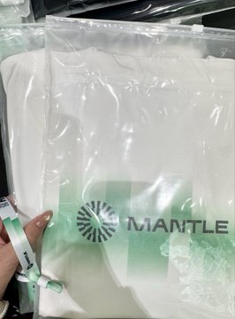 A hand holds a sealed clear plastic bag containing a white item with the "MANTLE" logo, likely event merchandise from the Mantle RWA Day. The image connects to the post as swag received at the blockchain conference, complementing the description of networking with other builders at the offline event.