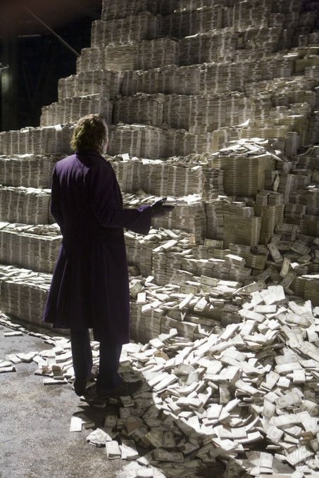 A photograph of a person in a purple coat standing before a massive, crumbling pyramid-shaped stack of bundled currency, with loose bills scattered across the floor.