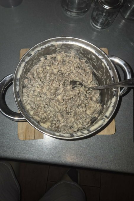 A stainless steel pot filled with cooked mushrooms or a similar dish on a wooden cutting board, with a spoon resting inside.
