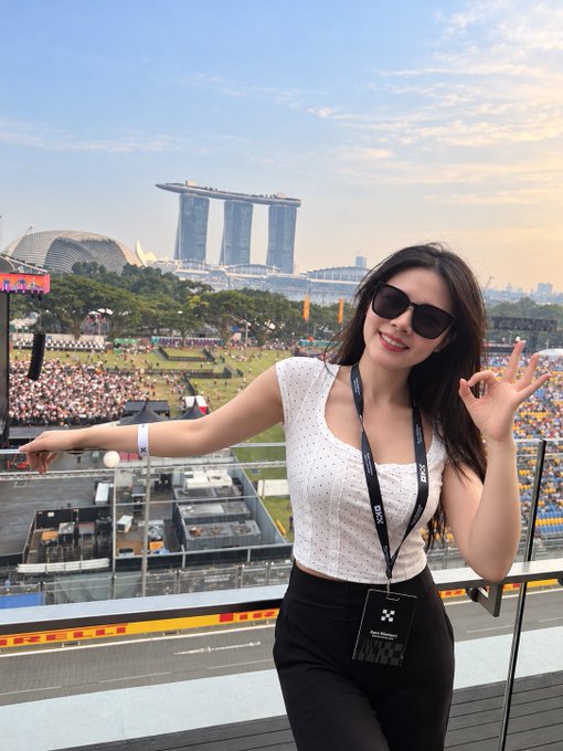 A woman wearing sunglasses and an OK gesture poses on a balcony overlooking the Marina Bay Sands in Singapore, wearing a 'Race Sponsor' lanyard.