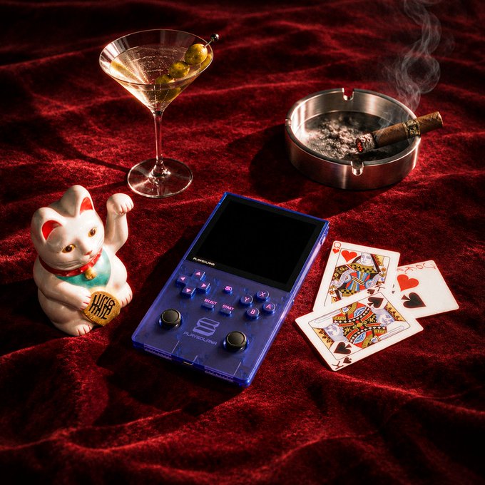 A translucent blue handheld gaming console sits on rich red velvet alongside a martini with olives, a smoking ashtray with a cigar, a lucky Maneki-neko cat figurine, and scattered playing cards (King of Hearts, Queen of Hearts, Jack of Spades), embodying the gambler's trappings of fortune—where bold bets meet chance and luck.