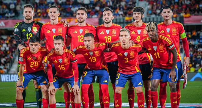 A Spain national football team poses for a group photo in their red kits, with visible jersey numbers including 13, 5, 12, 18, 4, 6 (back row) and 20, 16, 19, 3, 11 (front row). The image relates to the post about "La Roja," which is the nickname for the Spain national football team, as fans anticipate their match performance for a chance to win crypto giveaways.