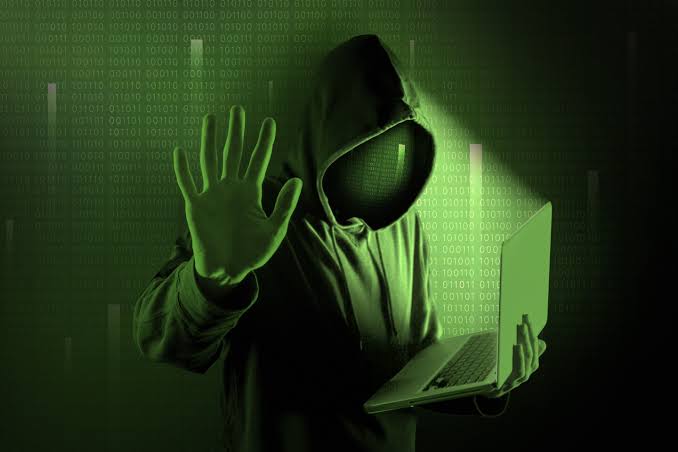 A stock image of a hooded hacker figure in green tones, holding a hand up in a stop gesture while using a laptop, with binary code overlaid in the background.