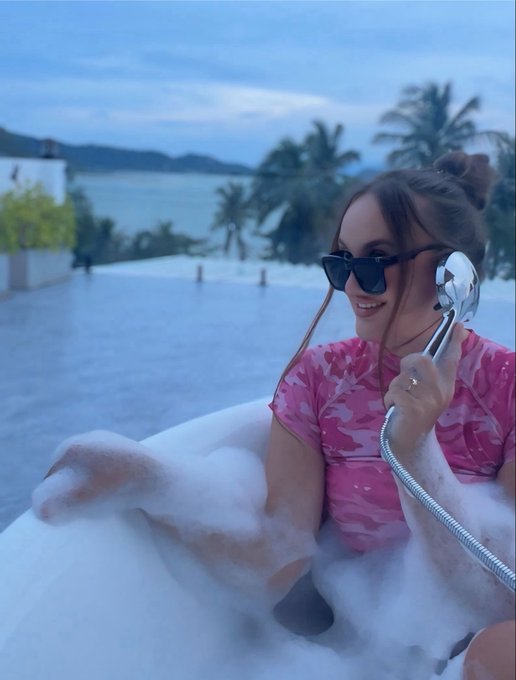 A woman wearing sunglasses and a pink camouflage-patterned top relaxes in a bubble-filled bathtub while holding a silver showerhead, with palm trees and water visible in the background.