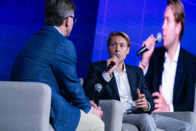 The image shows a group of people sitting around a table, engaged in conversation. One man is holding a microphone while talking with another person seated across from him. The third person is also present but not actively participating in the discussion. They are all dressed professionally, suggesting that they might be at an event or conference related to finance and value management.