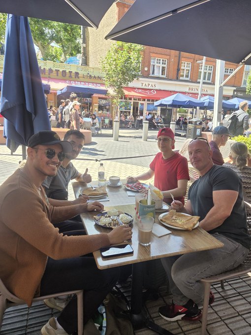 A group of four men sitting around a table outdoors at an outdoor café, with plates of food, drinks, and a busy pedestrian street with shops like 'COMPTOIR' and 'TAPAS' visible in the background.