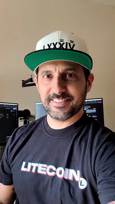 A smiling man wearing a "LITECOIN" branded t-shirt and a white snapback hat with green underbrim displaying "LXXXIV" (Roman numerals for 84), posing in what appears to be a tech or recording setup with multiple monitors in the background. The image ties directly to the post's thank-you message, showcasing the Litecoin merchandise received from @84Million alongside the related @84Mpodcast brand.
