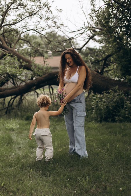 A woman in a white bikini top and baggy jeans smiles while handing a small bouquet of wildflowers to a young, shirtless toddler in white pants standing in a grassy backyard.