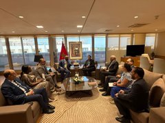 A formal meeting takes place in a high-rise office with several men in suits seated in a circle of beige armchairs around a low coffee table, with a Moroccan flag and a framed portrait visible in the background. The setting suggests an official delegation or business discussion in Morocco, aligning with the post's reference to IOTA's growing presence and partnerships in Africa.