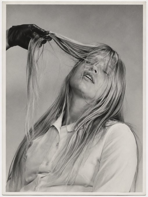 A black-and-white portrait of a blonde woman with long hair being pulled or lifted by a gloved hand, wearing a white collared shirt against a plain background.