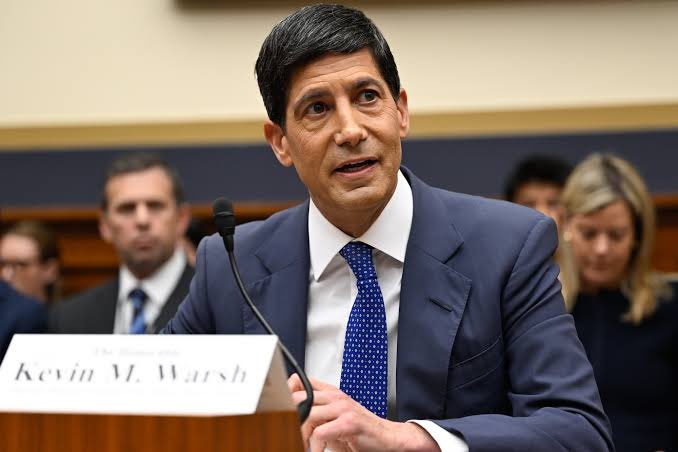 Kevin M. Warsh, identified by a nameplate, testifies at a congressional hearing, speaking into a microphone while wearing a blue suit and patterned tie.