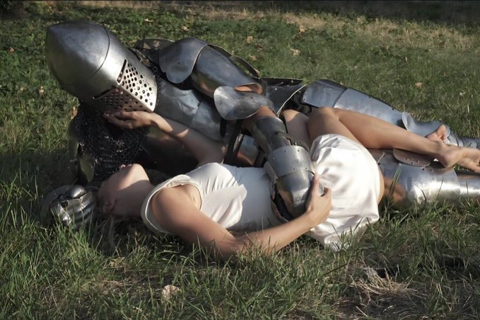 A knight in full metal armor lies on grass embracing a person dressed in white, in an artistic outdoor photograph.