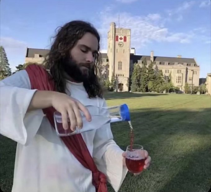 A man dressed as Jesus pours water from a clear bottle into a wine glass on a lawn in front of a large stone building with a tower.